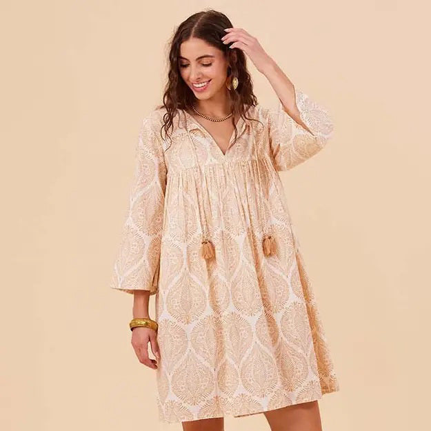 A woman smiles and looks down while wearing the ROCKFLOWERPAPER - ZAHARRA DRESS, a lightweight cream long-sleeve coverup with a subtle pattern and tassels, standing against a beige background.