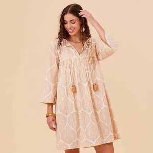 A woman smiles and looks down while wearing the ROCKFLOWERPAPER - ZAHARRA DRESS, a lightweight cream long-sleeve coverup with a subtle pattern and tassels, standing against a beige background.