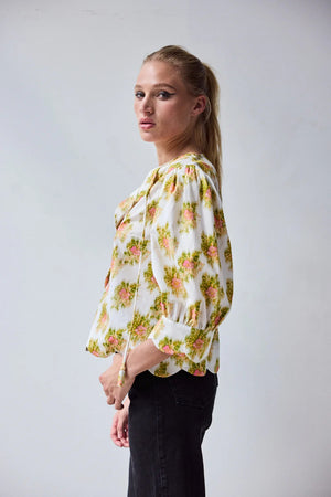 A woman with blonde hair in a ponytail wears the HOPE FOR FLOWERS Scalloped Shirt in Pink Warped Ikat by HOPE FOR FLOWERS and black high-waisted jeans, standing against a plain light background with her hands on her hips.