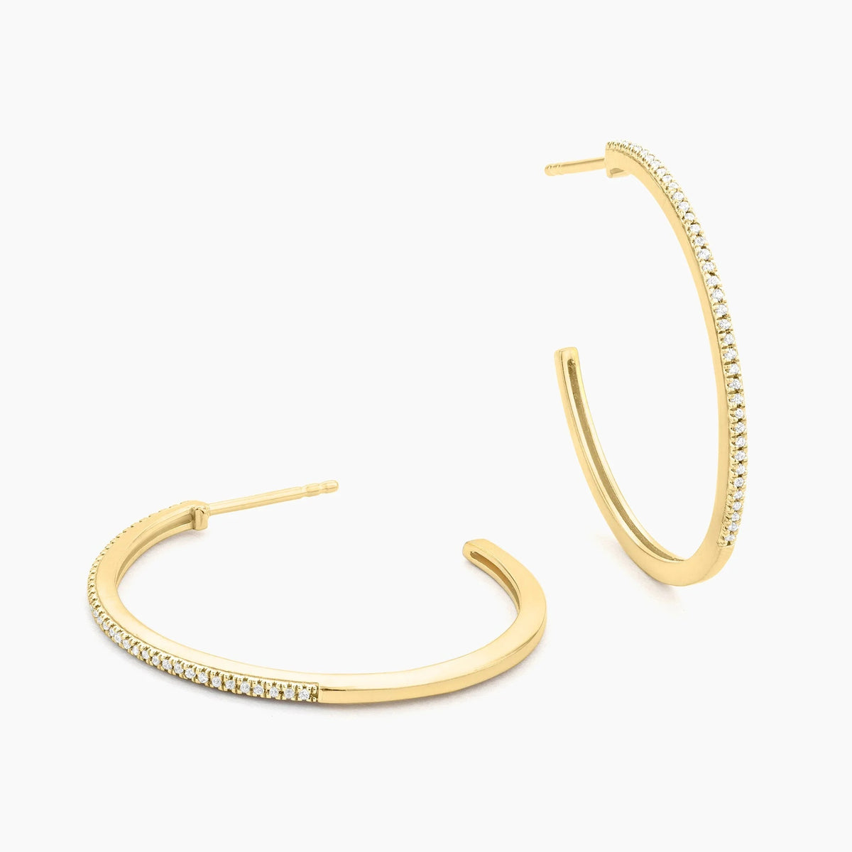 A woman with brown hair in a low bun is shown in profile against a plain light background, wearing the ELLA STEIN - A HOOP THING EARRINGS by ELLA STEIN—gold-plated hoops encrusted with small stones.