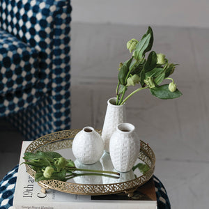 A gold tray displays the CREATIVE COOP EMBOSSED STONEWARE BASKET WEAVE VASE, SMALL, two white textured vases, and green foliage on a stack of books next to a blue patterned chair.