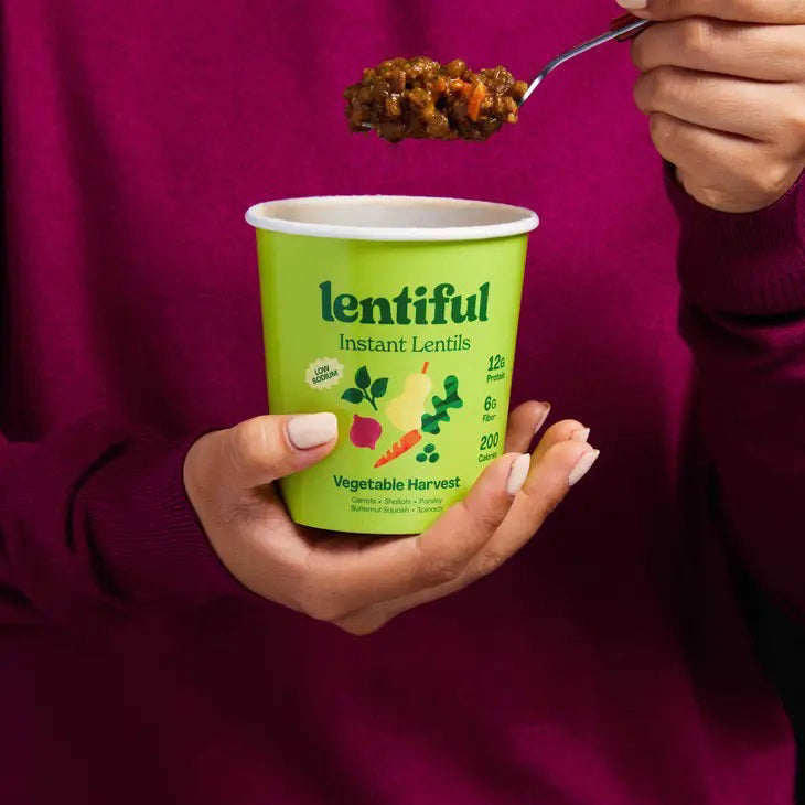 A container of LENTIFUL - LOW SODIUM VEGETABLE HARVEST INSTANT LENTILS by LENTIFUL sits on a yellow background, surrounded by fresh carrot, red onion, garlic, fennel, and squash.