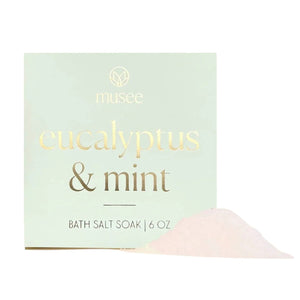 A square package labeled “MUSEE - EUCALYPTUS & MINT MINI BATH SALT SOAK” rests on a tray beside a green succulent, inviting you to enjoy the refreshing aroma of eucalyptus and mint with natural bath products from MUSEE.
