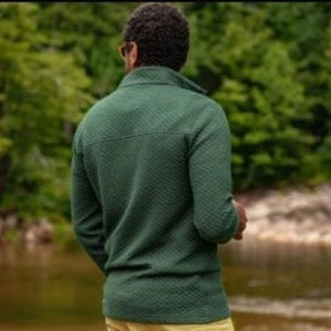 Wearing the CRIQUET Quilted Pullover in Heather Ivy, a person stands by a stone wall in khaki pants, holding a patterned mug and smiling, with greenery in the background—highlighting this classic-fit all-season layer from CRIQUET.