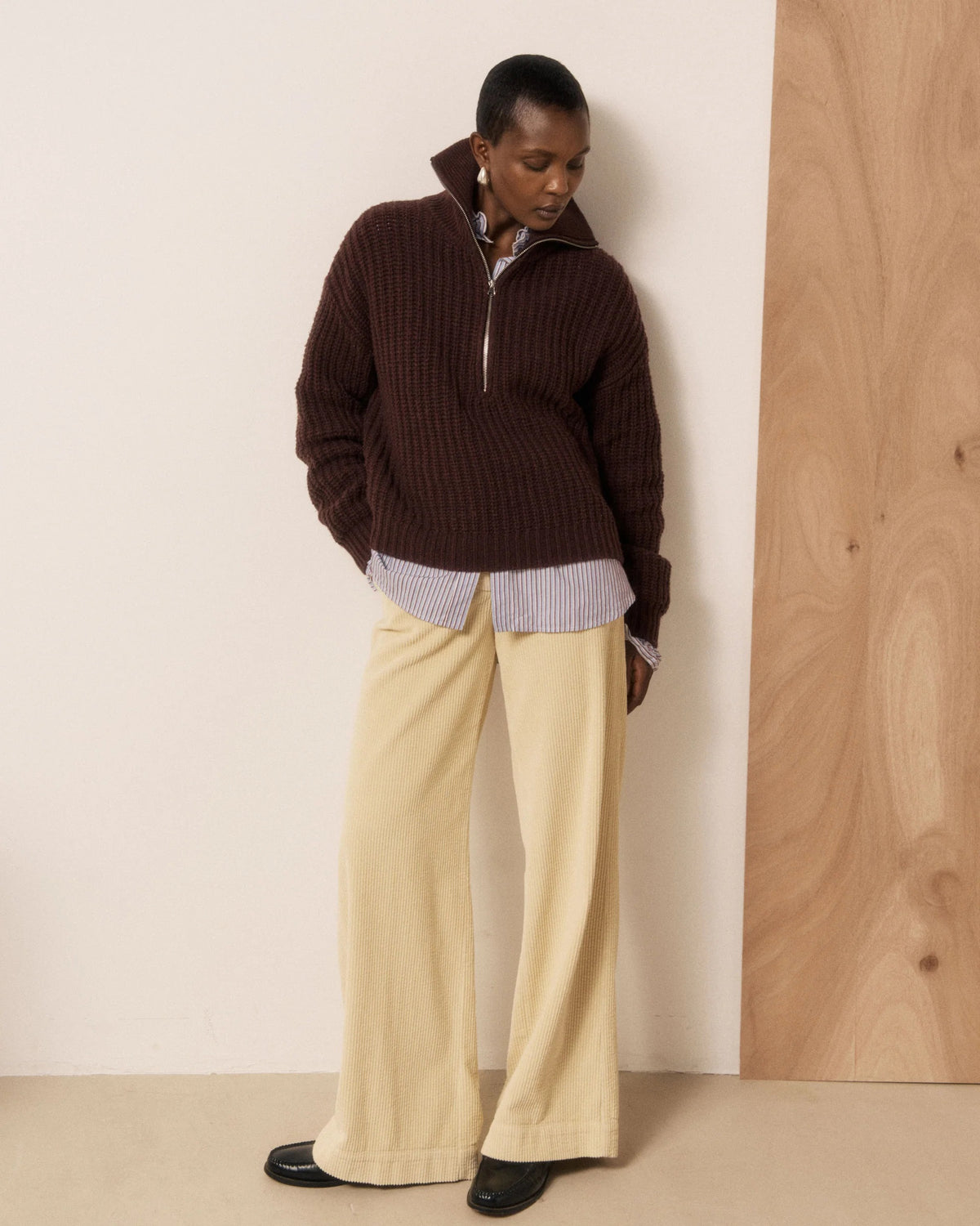 A person wearing the HARTFORD - MYPIO Sweater in Chocolate, styled over a striped shirt and paired with light-colored pants, stands in profile with hands in pockets against a neutral background.
