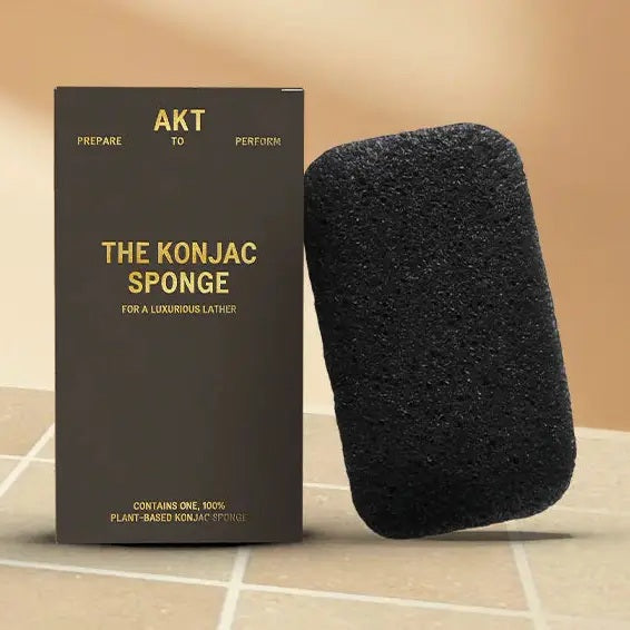 A black, rectangular AKT - KONJAC FACE AND BODY SPONGE by AKT LONDON, infused with activated charcoal, stands upright beside its packaging featuring the gold-embossed “AKT The Konjac Sponge” label on a brown background. Ideal for gentle exfoliation, this sponge leaves your skin feeling refreshed and smooth.