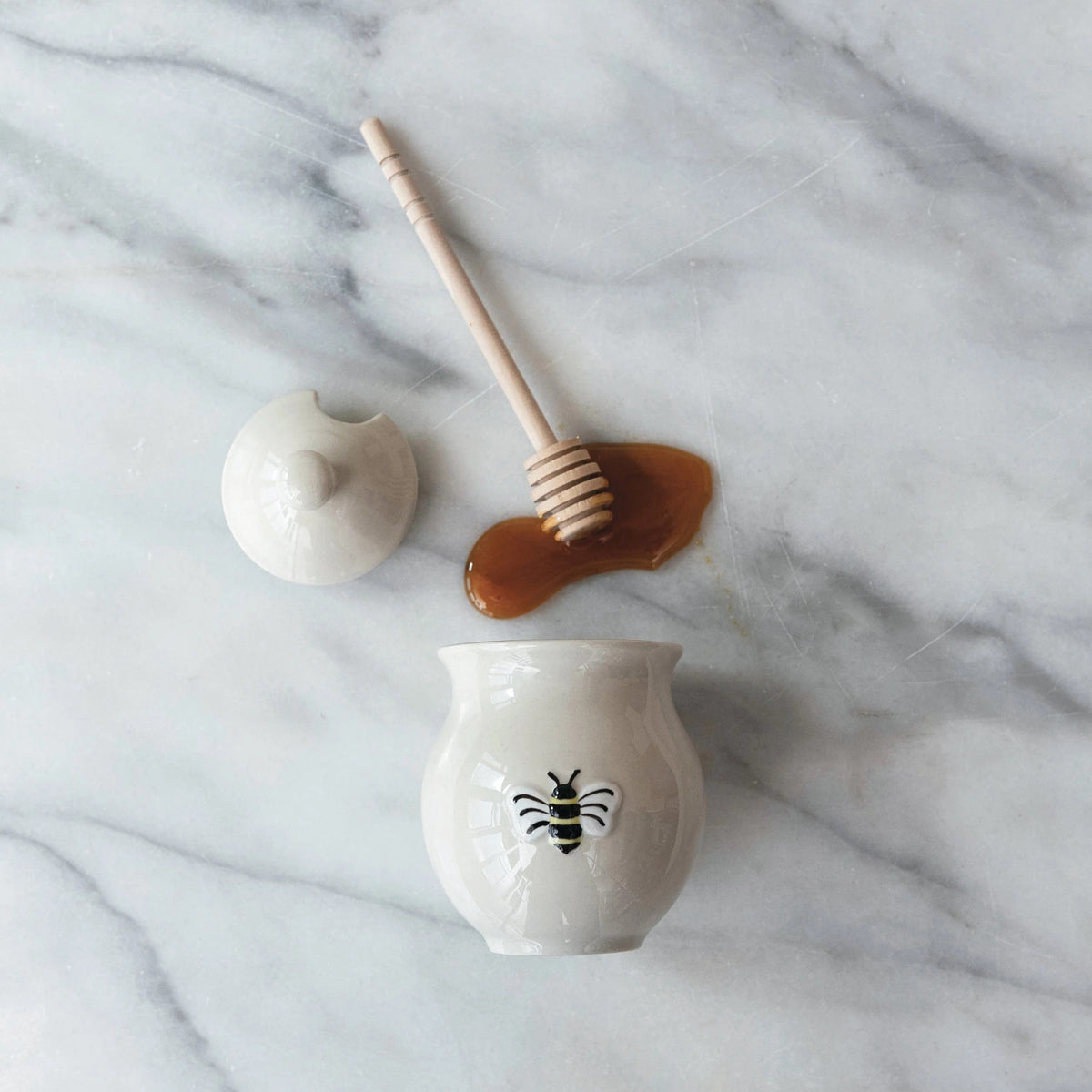 The HONEY JAR WITH EMBOSSED BEE AND WOOD DIPPER by CREATIVE COOP sits on a marble surface, partially covered by its lid. Honey oozes from the stoneware jar and pools beside it, with the wood dipper resting on top of the spill. The visually appealing setup conveys a sweet, natural theme.