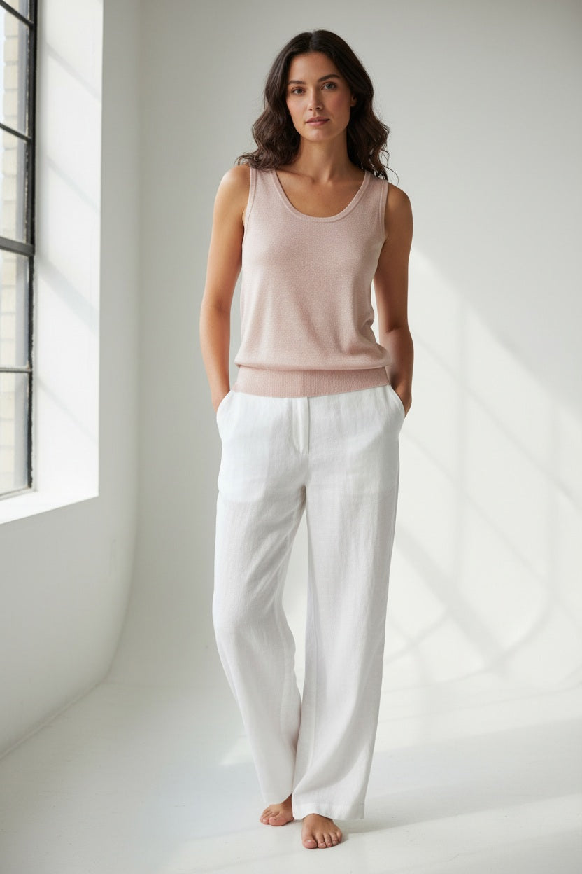 A woman with long dark hair models the KINROSS SLEEVELESS TANK IN LINEN, styled with white pants and hands in her pockets against a plain white background for a relaxed, everyday look.