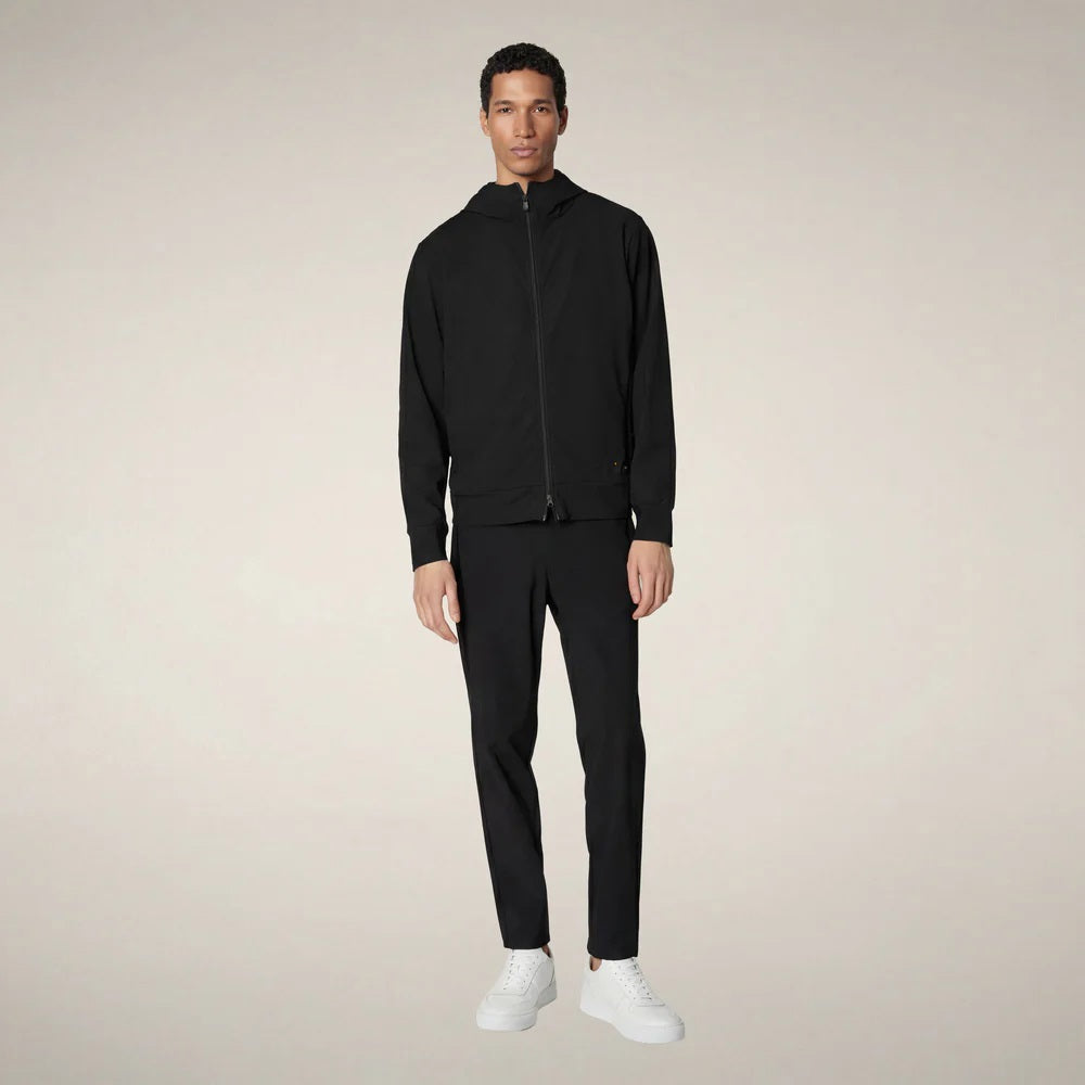 A man faces forward in the SAVE THE DUCK - LUIZ Hooded Jacket in Blue Black from SAVE THE DUCK, paired with black pants and white sneakers, set against a plain beige background.