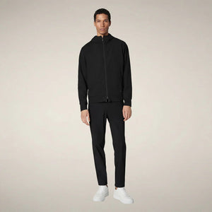 A man faces forward in the SAVE THE DUCK - LUIZ Hooded Jacket in Blue Black from SAVE THE DUCK, paired with black pants and white sneakers, set against a plain beige background.