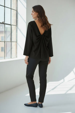 A person wears the MEIMEIJ TOP WITH KNOT BACK DETAIL in bright orange, paired with matching pants, standing against a plain light grey background. Their face is partially out of frame.