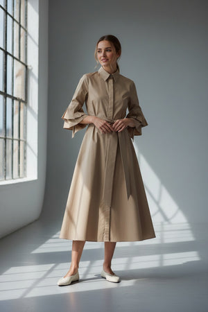 A woman in dark sunglasses and the MILVA MI - COTTON POPLIN DRESS WITH TIERED SCULPTURED SLEEVES by MILVA stands against a gray background, wearing flat woven shoes and sporting short hair.
