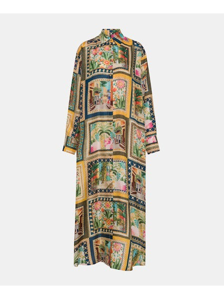 A person wears the MOMONI JAU DRESS, a colorful, artistic printed shirt dress, over blue high-waisted trousers against a plain backdrop. Brown sandals complete the effortless style as they gaze sideways.