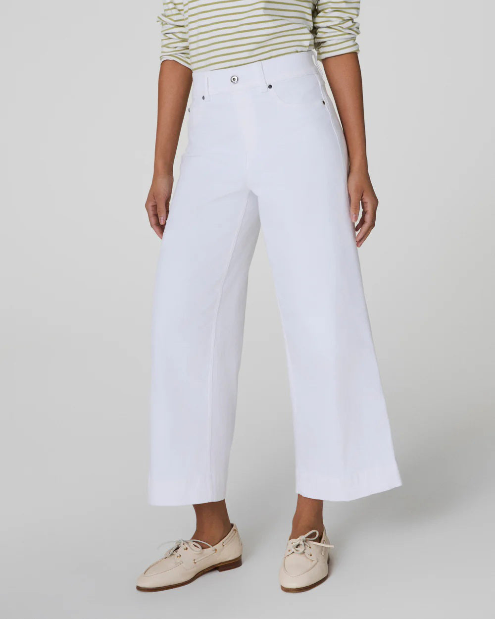 A person with short curly hair wears a striped long-sleeve shirt and high-waisted white SPANX PULL ON CROPPED WIDE LEG JEAN with TummyTamer Tech™. Against a plain background, they&#39;re holding a woven handbag and wearing beige shoes, exuding relaxed elegance.