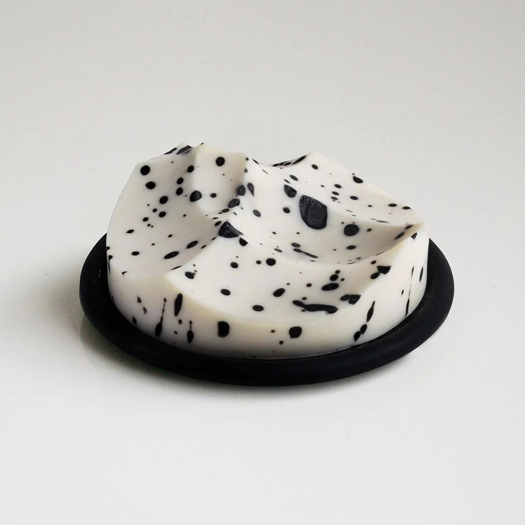 The UME STUDIO UME - WHITE GRAPEFRUIT ERODE SOAP, a round white handmade bar with black speckles, rests in a black foam-lined box beside its white packaging, both displayed on a silver surface with water droplets.