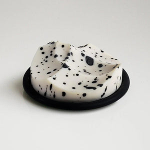 The UME STUDIO UME - WHITE GRAPEFRUIT ERODE SOAP, a round white handmade bar with black speckles, rests in a black foam-lined box beside its white packaging, both displayed on a silver surface with water droplets.