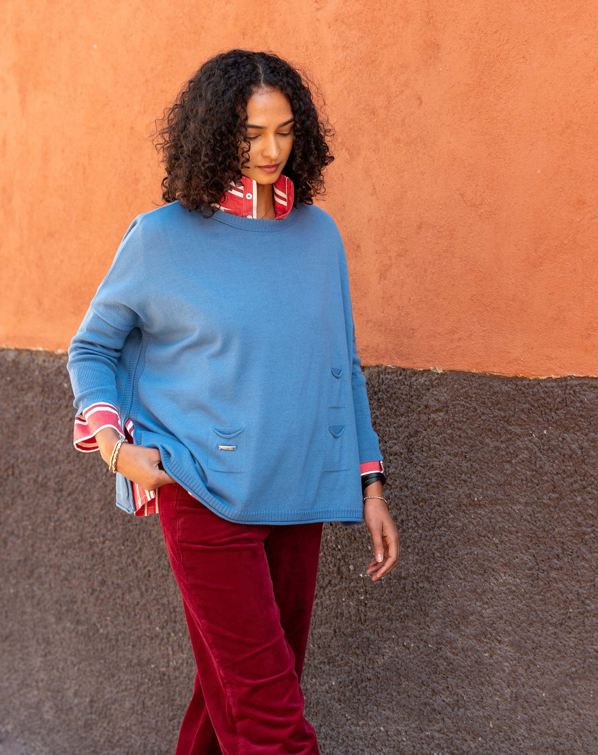 A person with curly hair walks along a two-toned wall, wearing the MERSEA - CATALINA CREWNECK SWEATER IN BLUE DUSK by MERSEA over a red collared shirt and red pants, looking down with a calm expression.