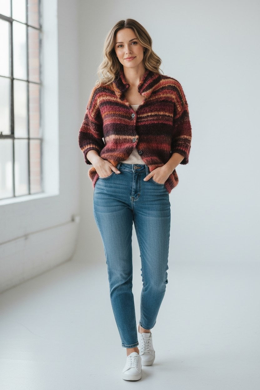 A woman stands indoors near a large window, wearing the FLOOR Painted Yarn Cardigan with 3/4 Sleeves—an oversized, chunky knit sweater in red and brown tones—paired with blue jeans and white sneakers. Her wavy hair and relaxed, confident expression complete the look.