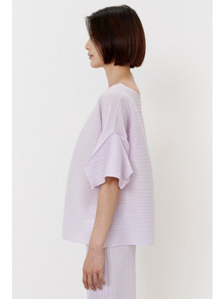A person wears the MEIMEIJ - PLISSE TOP, a loose-fitting, light lavender lilac top with wide sleeves, against a plain white background. Their minimalist ensemble is paired with matching pleated pants, complementing their dark shoulder-length hair.
