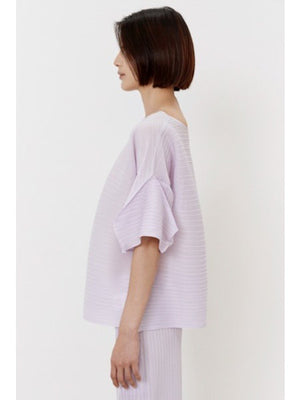 A person wears the MEIMEIJ - PLISSE TOP, a loose-fitting, light lavender lilac top with wide sleeves, against a plain white background. Their minimalist ensemble is paired with matching pleated pants, complementing their dark shoulder-length hair.
