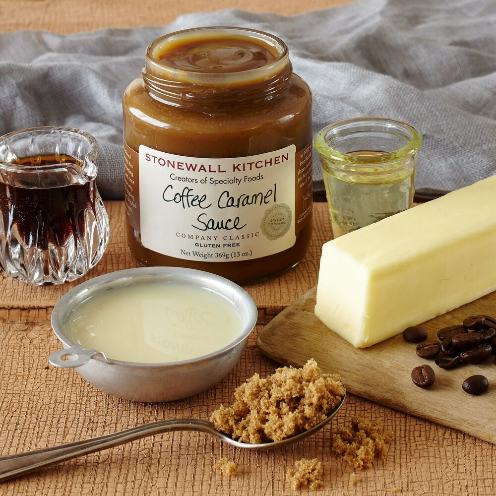A jar of STONEWALL KITCHEN - COFFEE CARAMEL SAUCE is displayed alongside a stick of butter, brown sugar, coffee beans, a glass of syrup, a small glass of oil, and a metal cup of cream on a wooden surface. This decadent caramel sauce from STONEWALL KITCHEN provides a rich and buttery sweetness that’s perfect for elevating your favorite desserts.