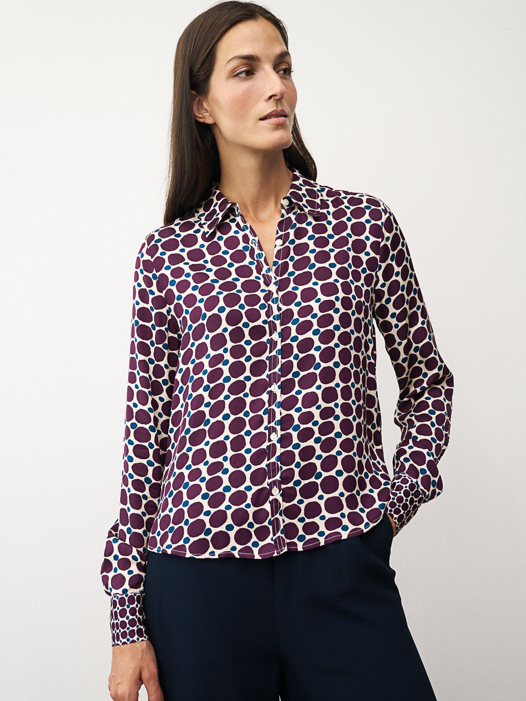 A person with straight brown hair adjusts the collar of the 0039 ITALY SIA BLOUSE, which features a geometric circle print with large green spots and small burgundy dots on cream. The side view highlights the blouse&#39;s viscose sleeve.