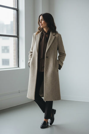 A sophisticated, single-breasted coat designed in a macro herringbone pattern on a white background. This SEVENTY coat, known as the SEVENTY - SINGLE BREASTED LUREZ MACRO HERRINGBONE COAT, features a notched lapel collar, long sleeves, and two buttons for closure. It’s crafted in light grey and white hues and proudly made in Italy with a label inside the collar that reads "Sustain.