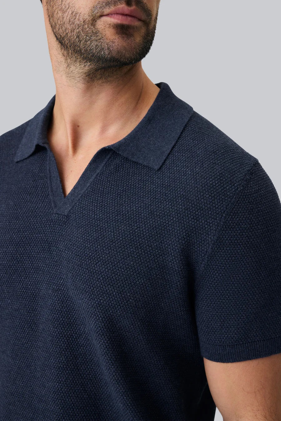 A close-up of a person’s arm and torso wearing the MACK WELDON Tech Linen Johnny Collar Polo Sweater in Navy Heather, paired with light gray pants featuring a buttoned pocket.