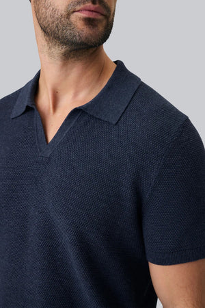 A close-up of a person’s arm and torso wearing the MACK WELDON Tech Linen Johnny Collar Polo Sweater in Navy Heather, paired with light gray pants featuring a buttoned pocket.