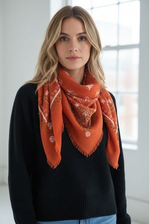 The STORIATIPIC - NELLY WOOL BANDANA SCARF by STORIATIPIC is an orange wool accessory measuring 100x100 cm, featuring fringed edges and adorned with white and pink floral embroidery accented with green details. Elegantly arranged in a loose loop on a white background, this scarf offers both style and comfort.