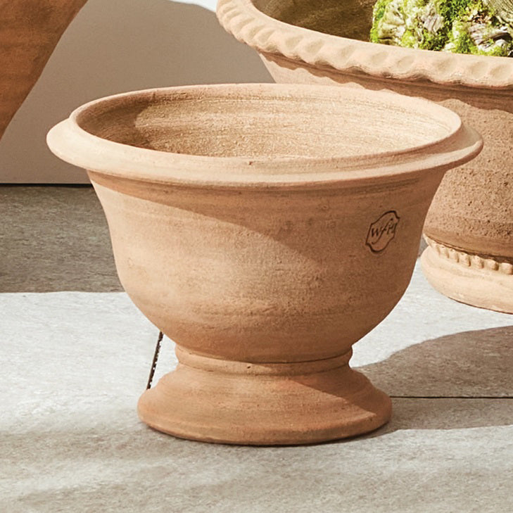 The NAPA HOME AND GARDEN - WAKEFIELD HANDMADE ASTON POT, crafted from aged terracotta with a wide rim and round base, sits on a stone surface beside another decorative planter in the background.