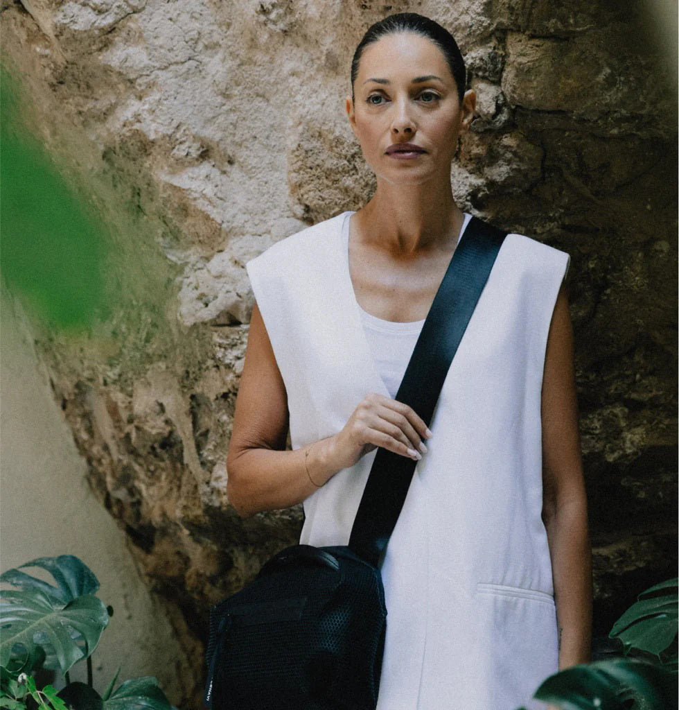 A person in a light beige suit carries the DANIELLA LEHAVI - BONNIE TRAVEL CROSSBODY, a large brown vegan bag with black zipper and strap, shown from shoulders to mid-thigh against a plain background.
