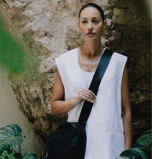 A person in a light beige suit carries the DANIELLA LEHAVI - BONNIE TRAVEL CROSSBODY, a large brown vegan bag with black zipper and strap, shown from shoulders to mid-thigh against a plain background.