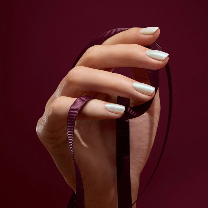 A person with light green painted nails holds a bottle of MANUCURIST INC’s GREEN™ Nail Polish in Fairy, wearing a purple knit top adorned with rhinestone embellishments.