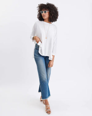 A woman with long, straight hair stands against a white wall, wearing the MERSEA Catalina Slub Tee in blue paired with white pants. The midweight textured cotton tee is from MERSEA. She looks at the camera with a neutral expression.