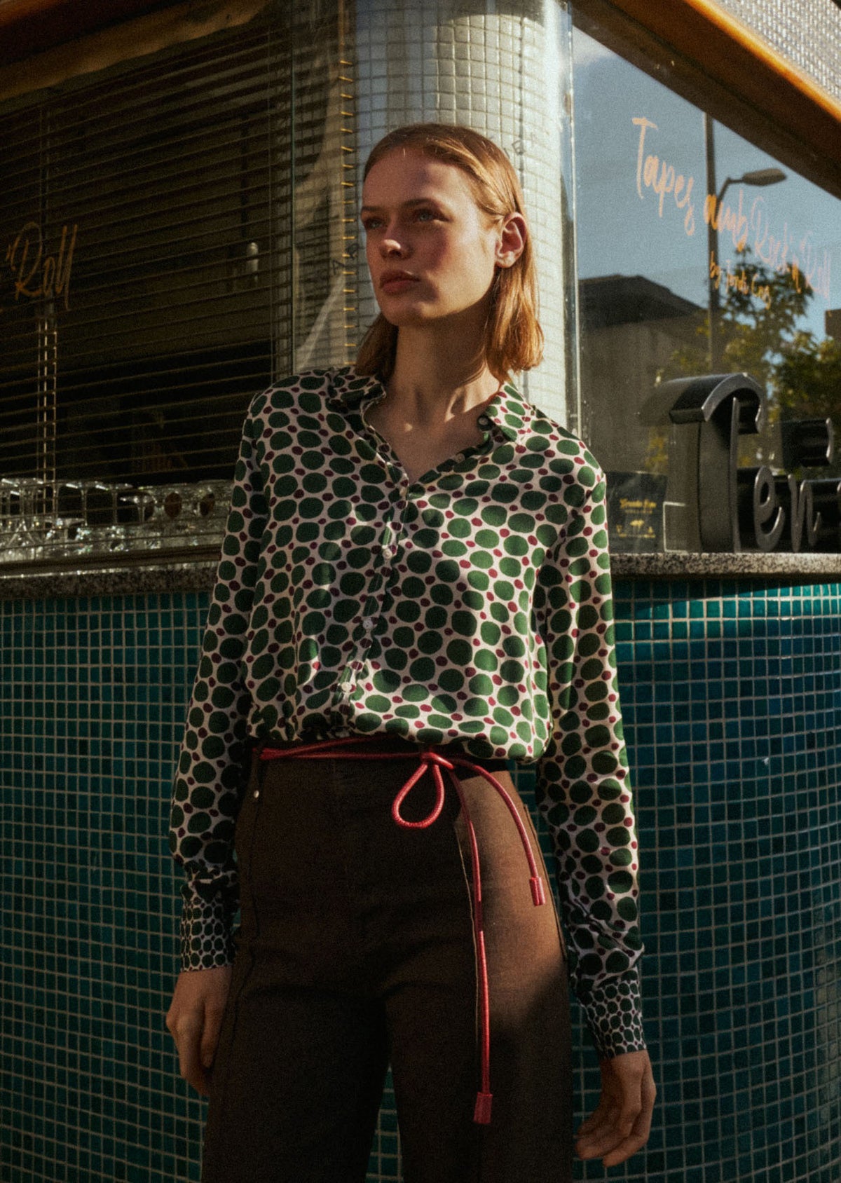 A person with straight brown hair adjusts the collar of the 0039 ITALY SIA BLOUSE, which features a geometric circle print with large green spots and small burgundy dots on cream. The side view highlights the blouse&#39;s viscose sleeve.