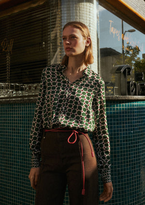 A person with straight brown hair adjusts the collar of the 0039 ITALY SIA BLOUSE, which features a geometric circle print with large green spots and small burgundy dots on cream. The side view highlights the blouse's viscose sleeve.