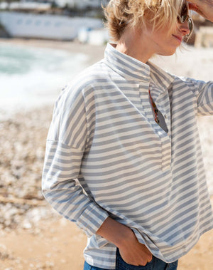 A person wearing sunglasses, denim shorts, and the MERSEA Emerson Pop Up Collar Top in Slate Blue Stripe stands on a rocky beach with buildings and trees in the background.