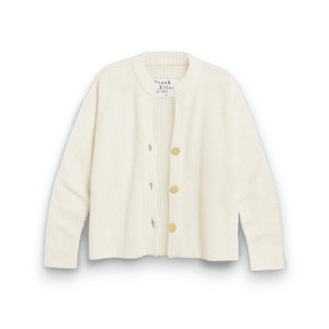 A woman with long hair, wearing a relaxed-fit FRANK & EILEEN MONTECITO CARDIGAN and beige shorts, stands outdoors smiling with a brown bag. A large plant pot and lush greenery enhance the serene scene.