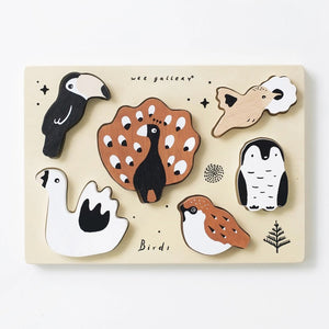 The WEE GALLERY - BIRDS TRAY PUZZLE is a wooden board featuring six bird-shaped pieces, including a toucan, peacock, penguin, swan, sparrow, and a small bird on a branch. This educational puzzle offers fun with its simple colors and shapes. The board is labeled with "wee gallery" and "Birds".