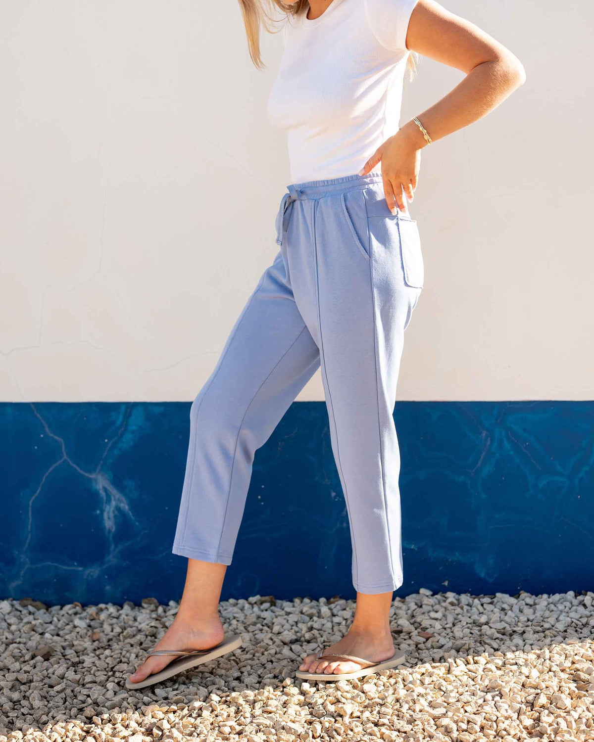 A person wears the MERSEA - FRENCHIE CIGARETTE PANT by MERSEA, paired with a white top and gray sneakers, standing on sandy beach ground with ocean waves and blue sky behind them, hands in their pockets.