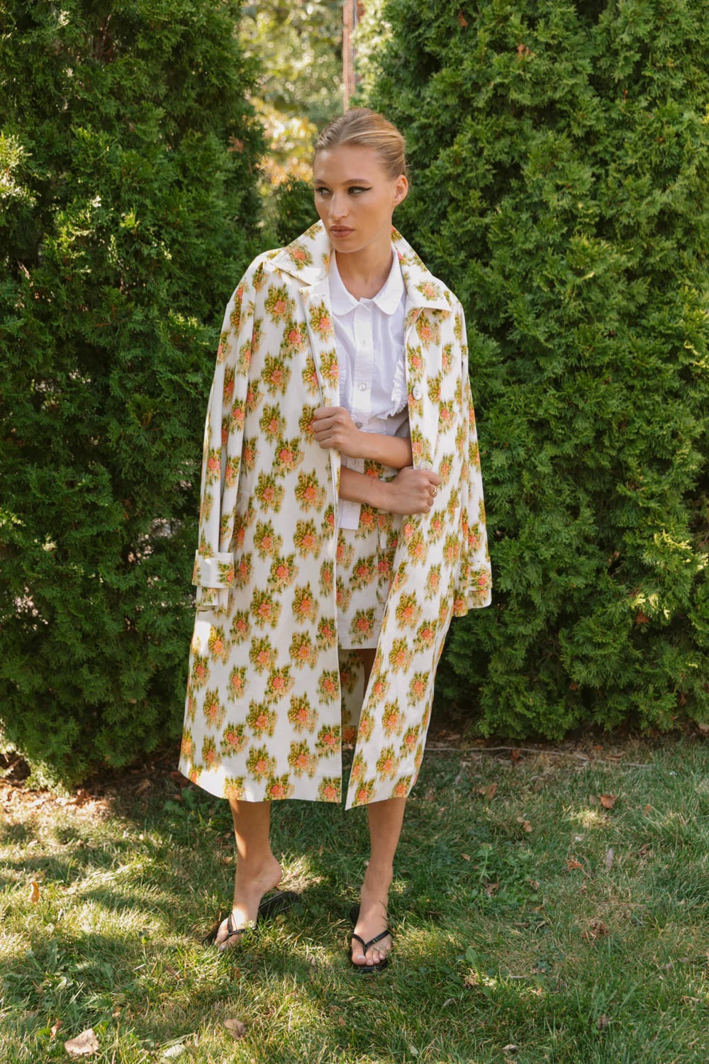 A person stands on grass before green bushes, wearing HOPE FOR FLOWERS' pink Warped Ikat coat over a white shirt and matching shorts with black sandals, looking to the side and highlighting the brand's commitment to ethical fashion.