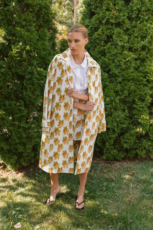 A person stands on grass before green bushes, wearing HOPE FOR FLOWERS' pink Warped Ikat coat over a white shirt and matching shorts with black sandals, looking to the side and highlighting the brand's commitment to ethical fashion.