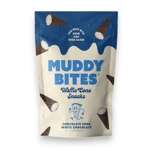 A person dressed in a light blue outfit sits on a plush, royal blue rug holding a bag of Muddy Bites Cookies & Cream Waffle Cones in one hand and a small white chocolate waffle cone snack in the other. The beige and blue bag features an illustration of chocolate-dipped waffle cones.