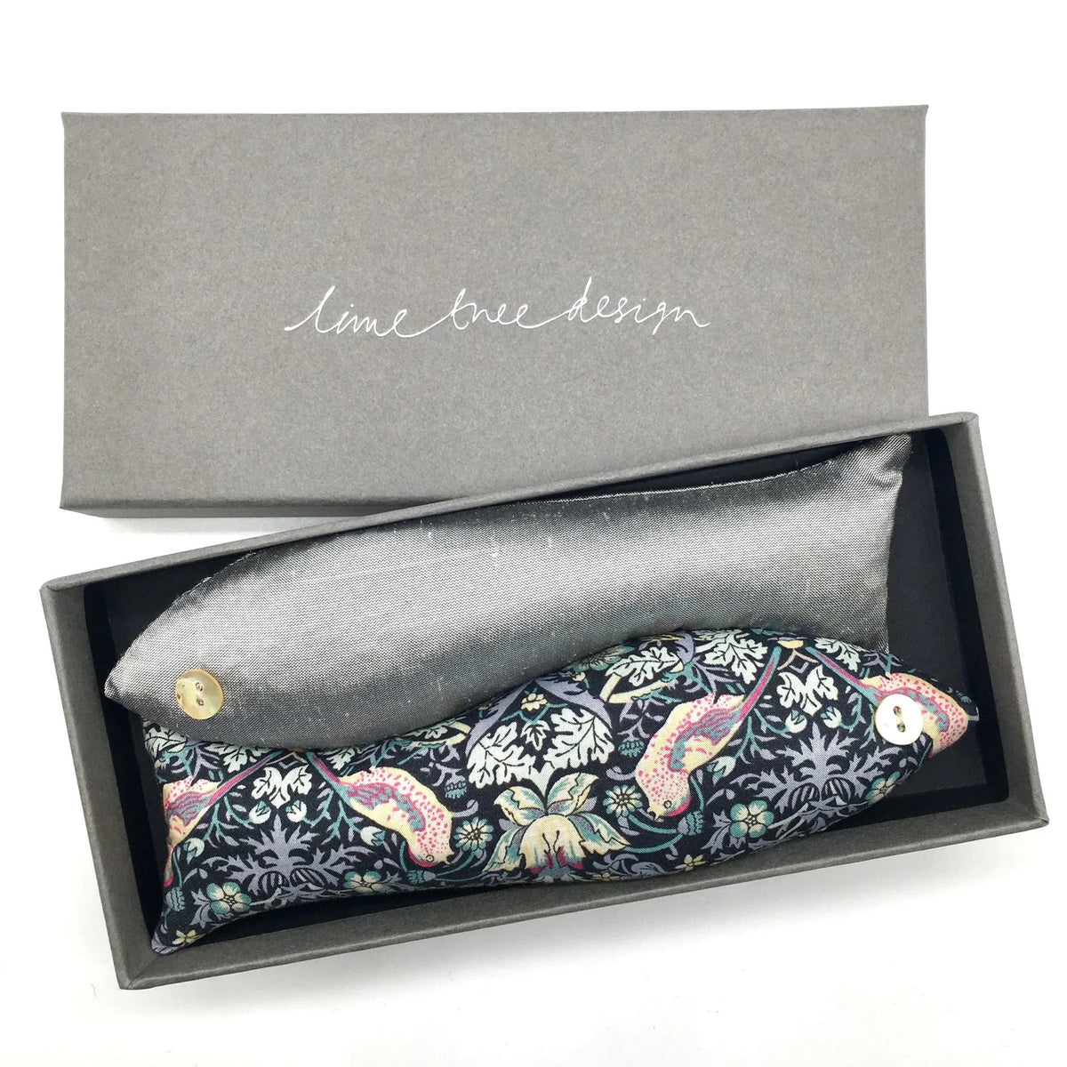 An open grey box with the text &quot;Lime Tree Collection Ltd&quot; on the lid contains two eye pillows. One pillow is grey, and the other is adorned with a colorful floral pattern made from Liberty Tana Lawn fabric. Both pillows have buttons at one end.