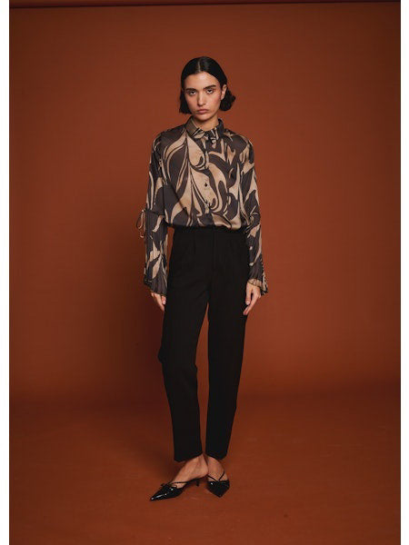 A woman poses against a brown background wearing the MILVA MI black and gold flare sleeve blouse by MILVA, paired with high-waisted black pants. Her dark hair is styled in a low bun.