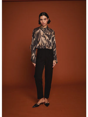 A woman poses against a brown background wearing the MILVA MI black and gold flare sleeve blouse by MILVA, paired with high-waisted black pants. Her dark hair is styled in a low bun.