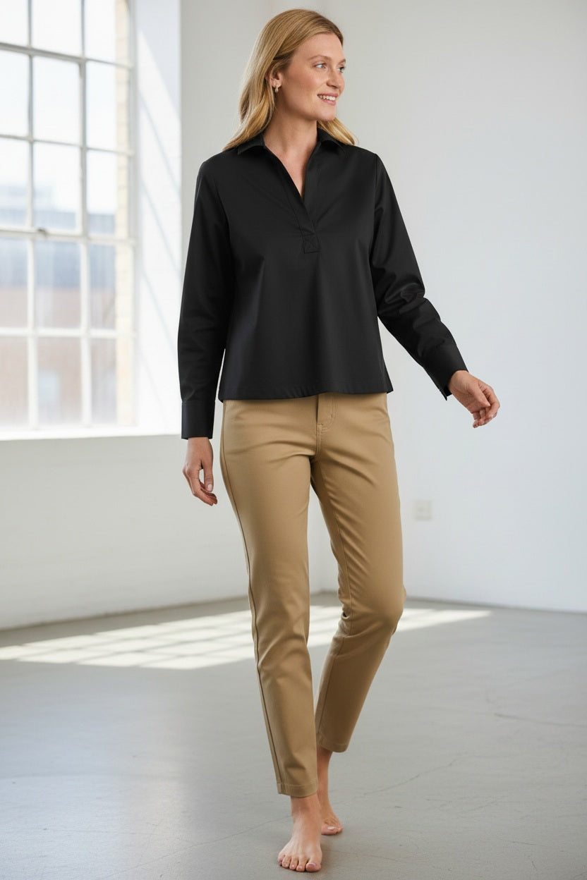 A woman stands barefoot indoors, smiling and looking to her left as natural light from a large window illuminates her effortlessly chic outfit. She wears the HINSON WU Monica Long Sleeve Popover with Buttons Down the Back, paired with tan stretch polished cotton pants, highlighting the relaxed fit of her ensemble.