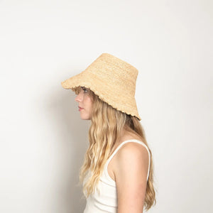 The WYETH Lacey Raffia Bucket Hat in Natural features a wide brim, textured woven pattern, and slightly scalloped edge. Displayed against a plain white background, this stylish hat is crafted by WYETH.