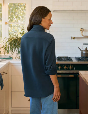 A woman with long brown hair, wearing the FRANK & EILEEN Eileen Relaxed Button Up Sweatshirt in Vintage Navy and light blue jeans, stands in a bright kitchen with white cabinets, a green stove, and a window filled with plants.